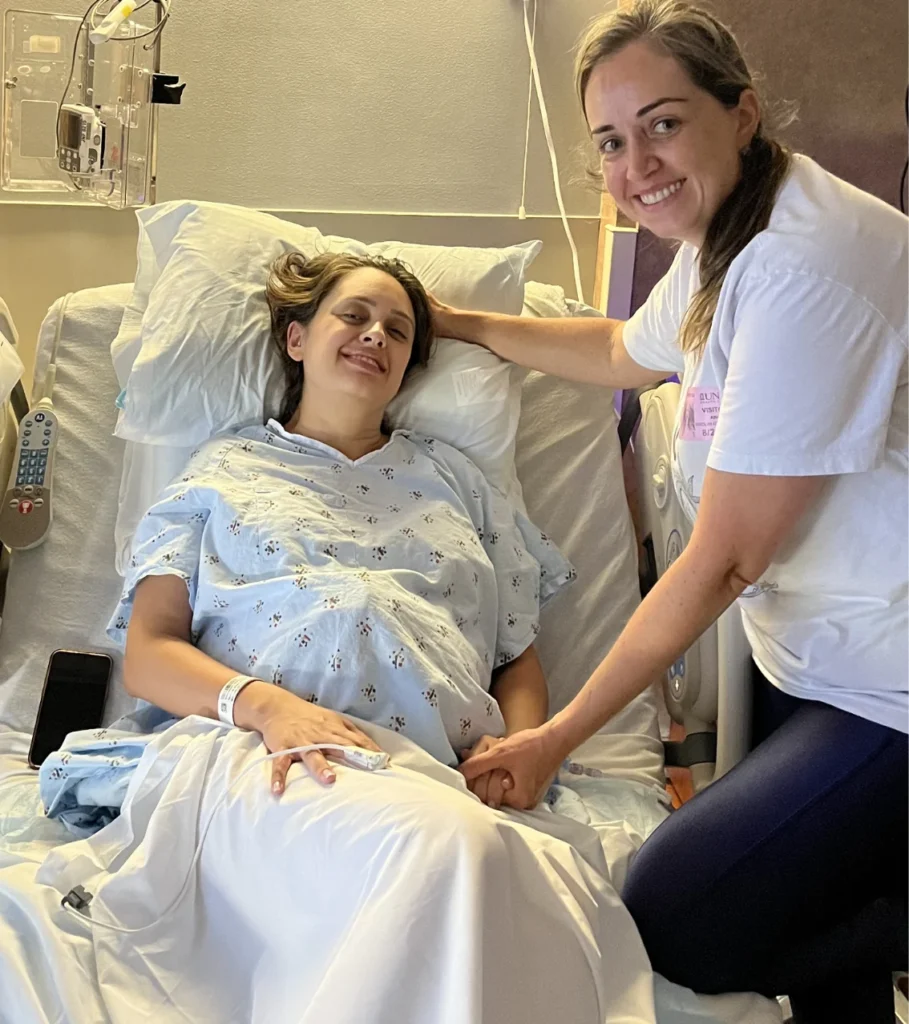 A smiling woman in a hospital bed holding hands with a woman in white standing beside her