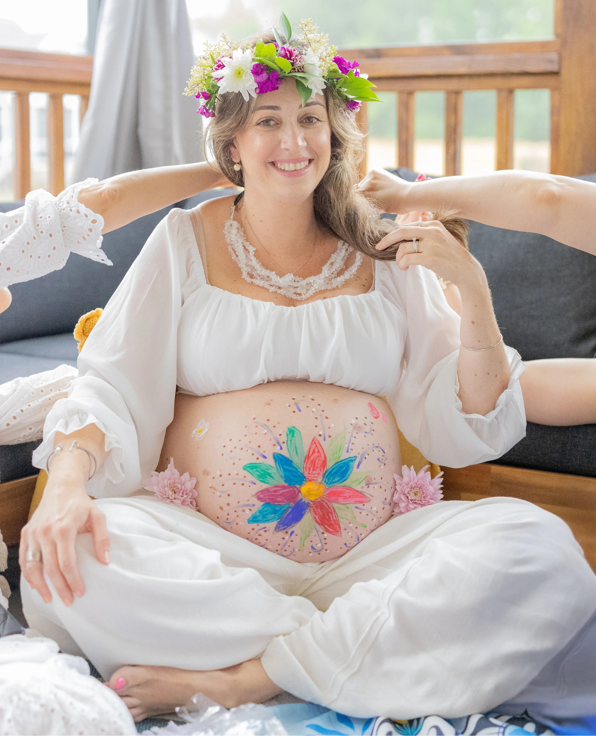 A woman in white sits cross-legged wearing a flower crown with a colorful flower painted on her belly